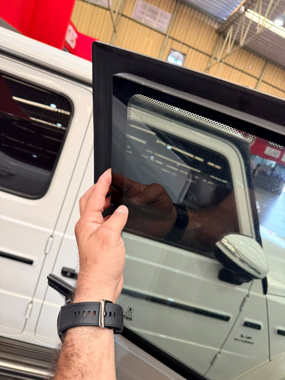 Mano sosteniendo un cristal blindado grueso y polarizado junto a una camioneta blindada blanca